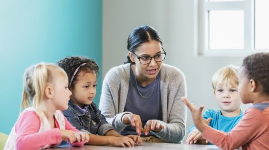 A multi-ethnic group of four preschool children with a mixed race African-American and Caucasian teacher, sitting around a table in a classroom.  The teacher, a young woman in her 20s, is looking at an African-American boy. The children are 4 years old.